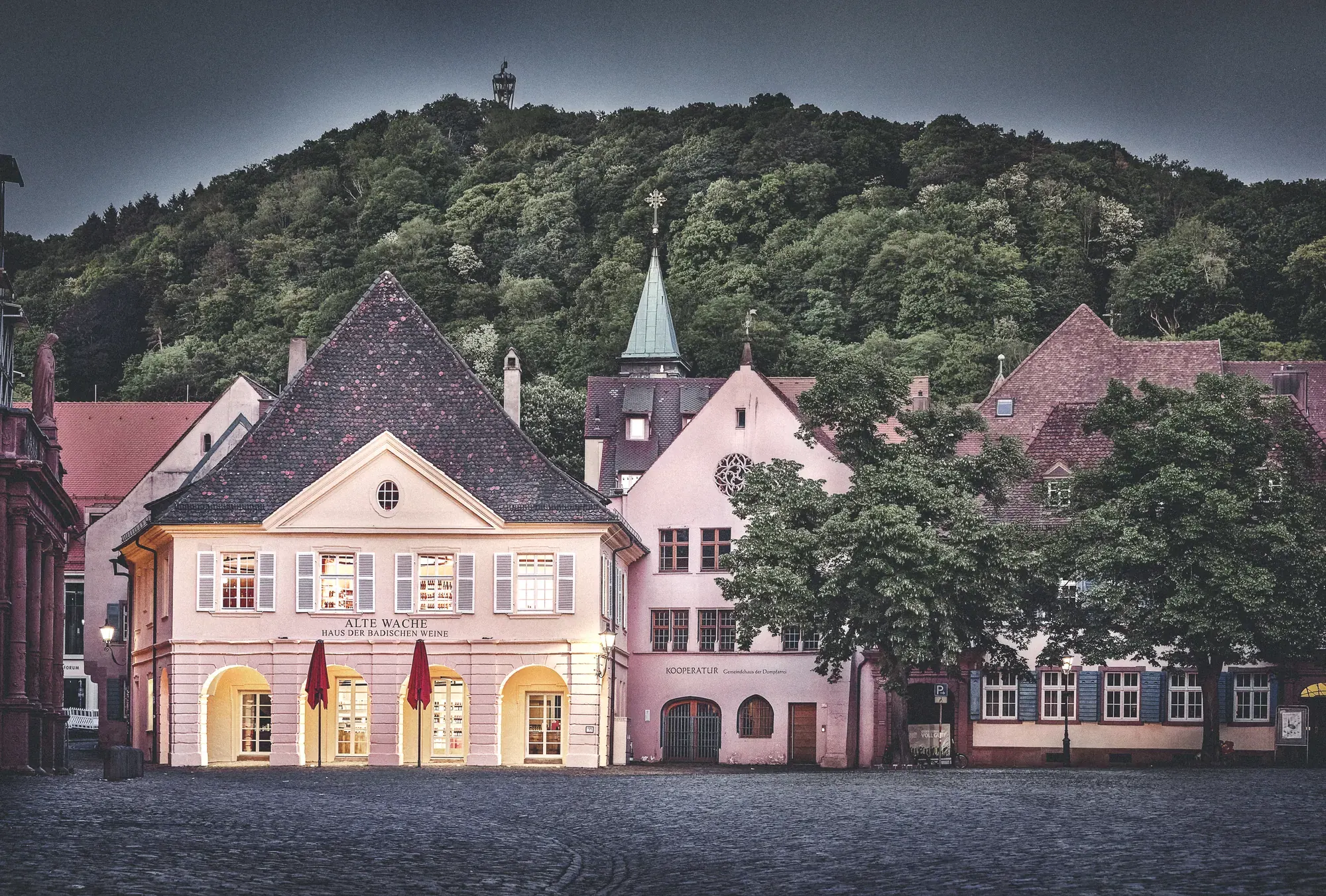 Alte Wache, Freiburg — historische Fassade am Münsterplatz in der Dämmerung, dahinter der Schlossberg