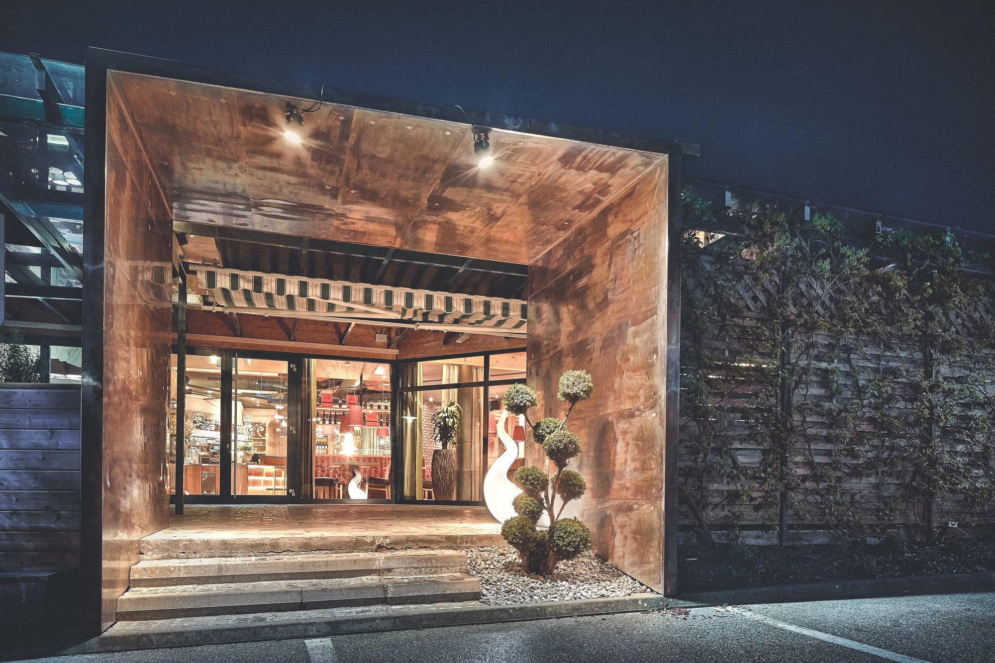 Restaurant Steiner, Pinsdorf — beleuchteter Eingang im Cortenstahl-Portal, davor Formgehölze im Kiesbett